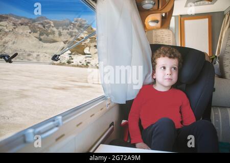 A small, curly-haired, Caucasian-looking boy, sitting by the open window of his parked motorhome with a spectacular view. Stock Photo