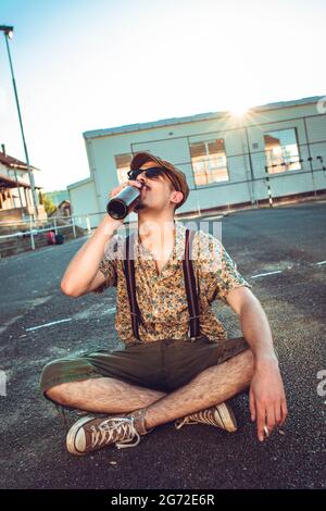 Young hipster man in retro clothing sits on the ground and drinking beer Stock Photo