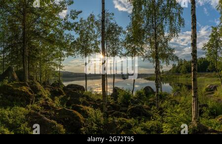 Landscape of moss covered boulders and birch trees on the shores of a calm idyllic lake in warm evening light and a sun star Stock Photo