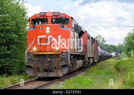 Saint John, NB, Canada - August 6, 2021: A Canadian National Railway train on an overcast say. Lights are on, trees and grass around tracks. Stock Photo