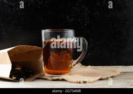 A glass mug of fragrant brewed flower tea on a wooden table, with a tea leaves in a paper bag. Selective focus. Stock Photo