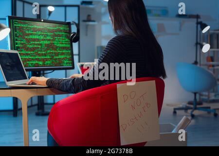 Female programmer working with computer and paper sheet with text DO NOT DISTURB in office at night Stock Photo