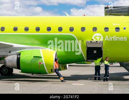 Airport personnel sits in an S7 airlines airplane jet engine, with his feet on ground, while workers working on fuselage. Stock Photo