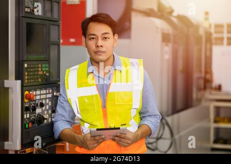 Smart adult professional Asian male engineer worker standing with CNC heavy machine and tablet computer in hand. Stock Photo