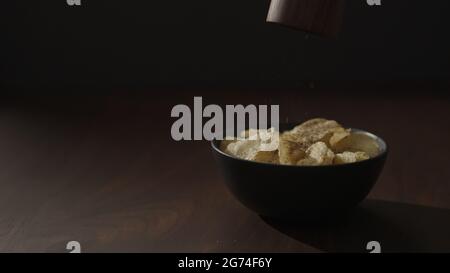 freshly ground pepper falling on organic potato chips in black bowl on walnut table, wide photo Stock Photo