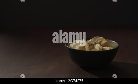 Slow motion freshly ground pepper falling on organic potato chips in black bowl on walnut table, 120fps Stock Photo