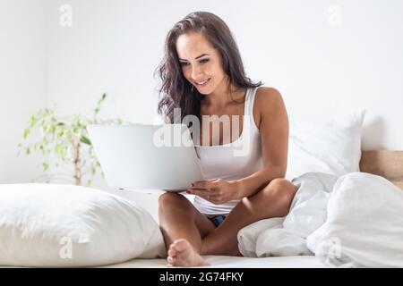 Woman works on her laptop sitting on a bed surrounded by white pillows. Stock Photo
