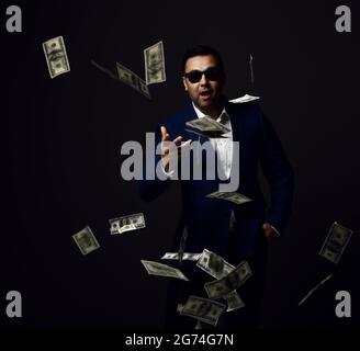 Portrait of rich successful businessman in blue jacket and sunglasses standing throwing dollars cash at camera