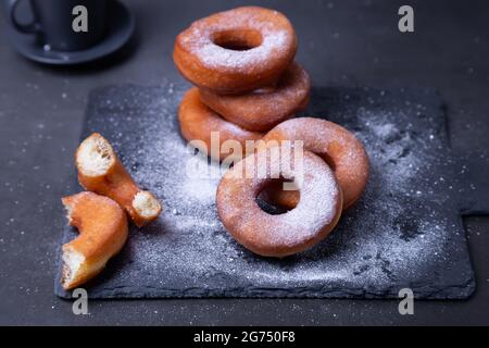 Donuts with powdered sugar and a cup of coffee. Traditional donuts in the shape of a ring fried in oil, on a black background. Junk food. Close-up. Stock Photo