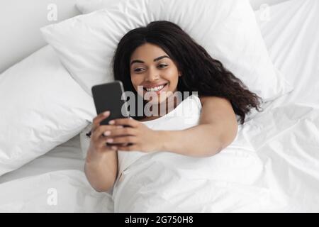 Happy young african american lady lying in bed and having video call on smartphone, lying in bed under white blanket Stock Photo