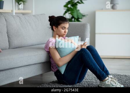 Stressed Indian teen girl sitting on floor with cellphone and reading bad message at home Stock Photo