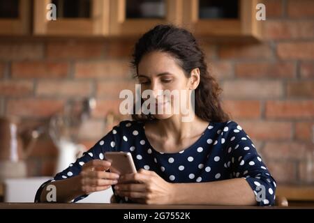 Focused Latin woman reading text on cellphone screen in kitchen Stock Photo