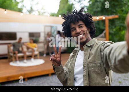 Black guy taking selfie near motorhome, gesturing peace, resting with his multiracial friends outdoors, enjoying summer Stock Photo