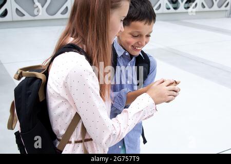 Two schoolchildren, boy and girl with backpacks behind their backs, found beetle in concrete courtyard campus, holding it in their hands Stock Photo