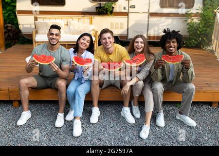 Happy diverse friends sitting near camper van with pieces of watermelon, eating tasty fruit, having fun summer vacation Stock Photo