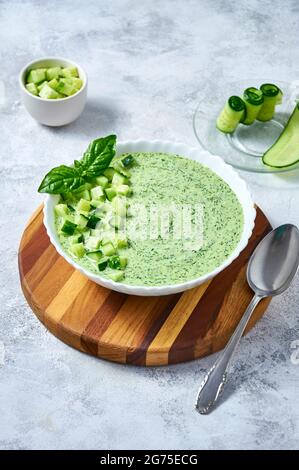 Cucumber Gazpacho - cold summer soup with basil in white bowl on wooden board on light background. Stock Photo