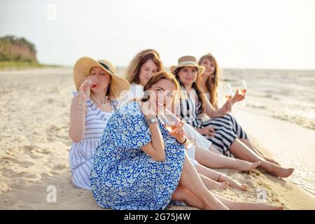 Group of delighted female friends enjoying wine and talking with each other while relaxing on sandy beach near waving sea in summer Stock Photo