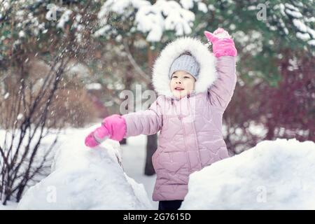 Smiling happy child girl in warm pink winter clothes throws a snowball in winter Stock Photo