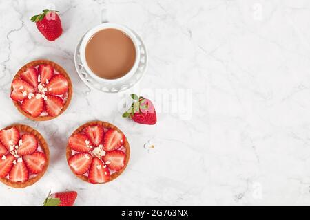 Small tartlet pastries topped with strawberry fruits and white chocolate sprinkles next to coffee cup and copy space Stock Photo