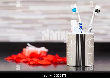 On a dark surface of a bathroom cabinet there is, in the foreground, a jar with two toothbrushes and a tube of toothpaste, in the background, out of f Stock Photo
