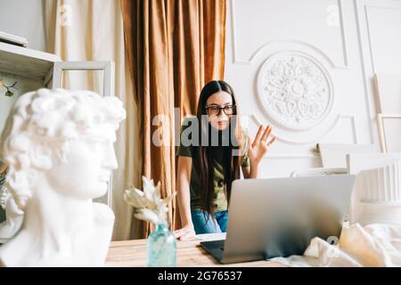Young caucasian woman in eyeglasses working on laptop computer in home art studio using video call... Stock Photo