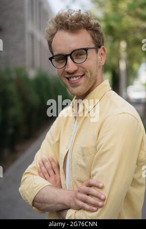 Portrait of handsome smiling man wearing yellow shirt and stylish eyeglasses looking at camera standing on the street. Successful business and career Stock Photo