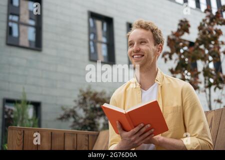 Young smiling student studying, exam preparation, reading book, sitting on bench in university campus Stock Photo
