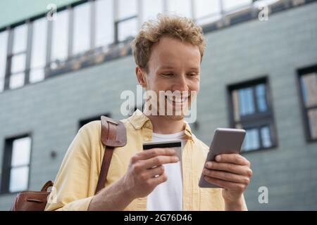 Handsome smiling businessman holding credit card, using smartphone for online shopping standing on the street. Young happy freelancer receive payment Stock Photo