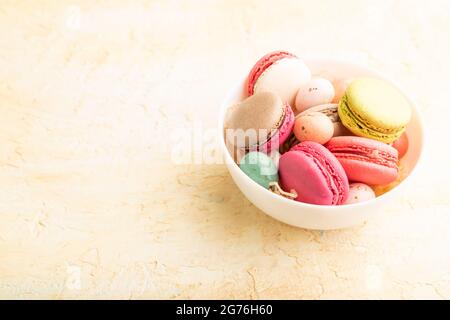Multicolored macaroons and chocolate eggs in ceramic bowl on beige concrete background. side view, copy space, still life. Breakfast, morning, concept Stock Photo