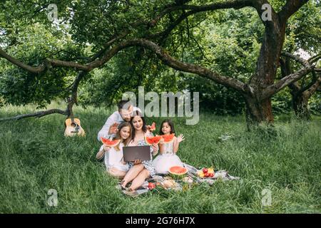 Young parents with two little daughters having video call on laptop during picnic time at green garden. Happy family talking with relatives online. Concept of people and technology. Stock Photo