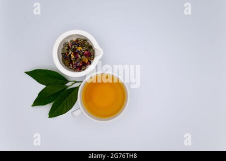 Dried herbs and flower petals in a white mortar, a cup of fresh tea and greenery against a white background Stock Photo