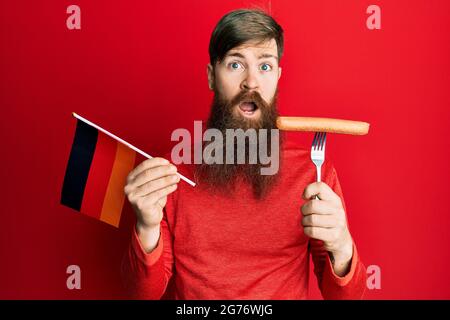 Redhead man with long beard holding fork with pork sausage and germany flag clueless and confused expression. doubt concept. Stock Photo