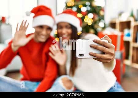 Young latin couple smiling happy having video call using smartphone sitting on the floor by christmas tree at home. Stock Photo