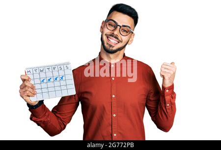 Young hispanic man with beard holding travel calendar pointing thumb up to the side smiling happy with open mouth Stock Photo