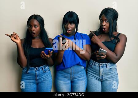 Three young african american friends using smartphone surprised pointing with finger to the side, open mouth amazed expression. Stock Photo