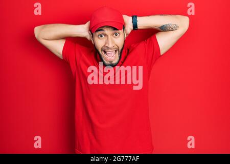 Hispanic man with beard wearing delivery uniform and cap crazy and scared with hands on head, afraid and surprised of shock with open mouth Stock Photo
