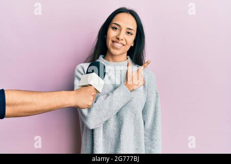 Young hispanic girl being interviewed by reporter holding microphone smiling cheerful pointing with hand and finger up to the side Stock Photo