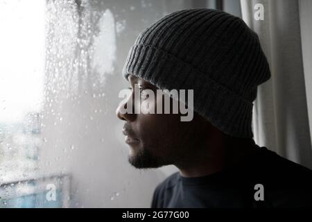 Portrait of a sad African American man looking out through a glass window on a wet cold rainy day, water droplets running down the window. Stock Photo