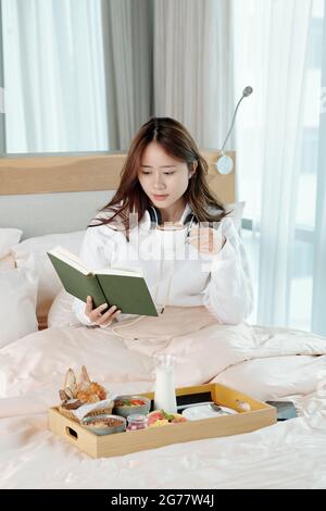 Pensive pretty young woman drinking cup of cappuccino and reading reading interesting book when sitting on bed at home Stock Photo