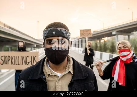 Portrait of serious brutal man in mask wearing headband and facial mask standing at street against protesting people with signs Stock Photo