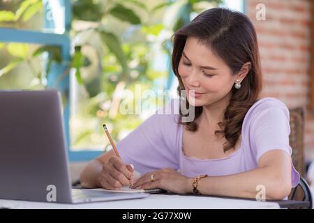 The beautiful middle-aged woman sitting in food shop and working with laptop note computer and writing on memo paper. Freelancer job and work from any Stock Photo