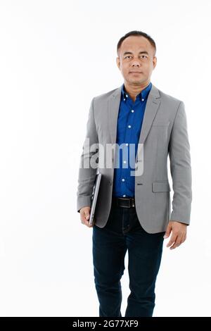 A self-confident middle age senior Asian man in a blue business casual shirt with a grey jacket standing and holding the notebook laptop computer and Stock Photo