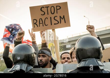 Group of displeased young American people raising fists and banner while asking to stop racism, police keeping crowd Stock Photo