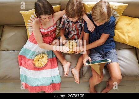 Kids eating potato chips when sitting on sofa and watching movie or animated cartoon on tablet computer, view from above Stock Photo