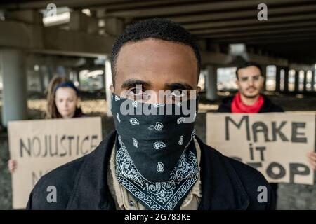 Portrait of young Black protester in bandana on face standing against people with signs under bridge Stock Photo