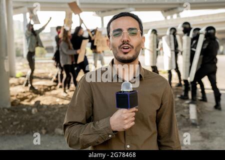 Portrait of young mixed race journalist with beard speaking into microphone while covering riot: protestors arguing with riot police in background