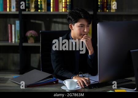 Young and handsome Asian businessman new generation in suit sitting in private working room and looking to computer screen with serious and thoughtful Stock Photo