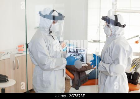 Dentist doctors in overall using tablet explaining dental x ray in stomatologic office during coronavirus. Man wearing face shield and mask showing to nusrse radiography using digital device Stock Photo
