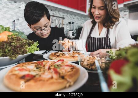 Asian mom and son sitting in home kitchen and eating homemade pizza together with various kinds of vegetables. Idea for happiness of good time sharing Stock Photo