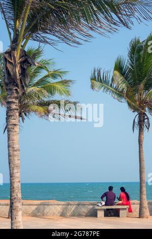 Indian couple sitting by promenade beneath palm trees overlooking blue sea and blue sky, Puducherry (Pondicherry), Tamil Nadu, India Stock Photo